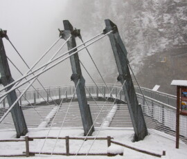 La passerella sospesa sovrastata dalle nubi sembra un essere alieno surreale. Il pannello e la parete rocciosa sul versante opposto, al di fuori delle nubi, danno profondità di campo e riportano l’intera struttura alla realtà
